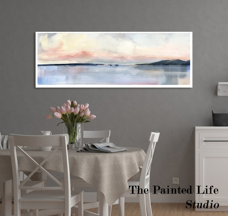 May include: A watercolor painting of a seascape with a pink and yellow sky, a blue body of water, and a dark green mountain range in the distance. The painting is framed and hanging on a gray wall above a white dining table with a white tablecloth and four white chairs. The table is decorated with a vase of pink tulips and a magazine. The text "The Painted Life Studio" is visible in the bottom right corner of the image.