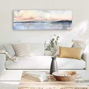 May include: A white sofa with a throw blanket and pillows in a living room setting. A painting of a pink and blue sunset over a body of water hangs on the wall above the sofa. The painting is titled "The Painted Life Studio".