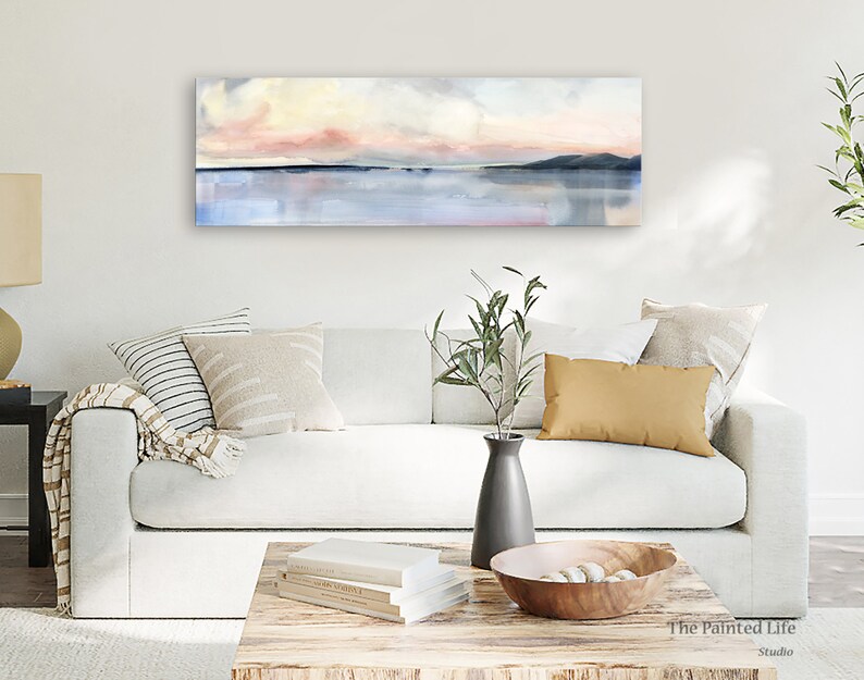May include: A white sofa with a throw blanket and pillows in a living room setting. A painting of a pink and blue sunset over a body of water hangs on the wall above the sofa. The painting is titled "The Painted Life Studio".