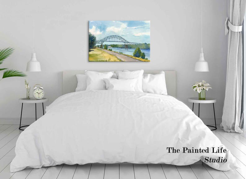 May include: Watercolor painting of a bridge over a river, with blue skies and green trees. The artwork is displayed above a bed with white bedding. The Painted Life Studio is printed on the bottom right.