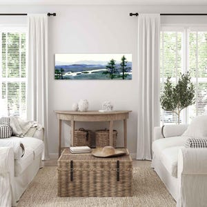 May include: A watercolor painting of a landscape with a lake and trees. The painting is framed and hanging on a white wall in a living room. The room is decorated in a farmhouse style with white furniture and wicker baskets.