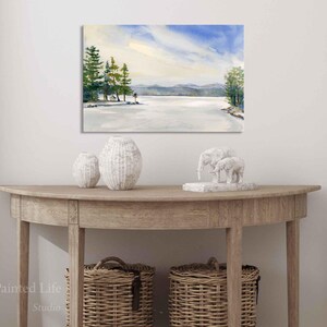 May include: A watercolor painting of a snowy lake scene with a group of trees on the shore. The sky is blue with white clouds and the water is a light blue. The painting is framed and hanging on a white wall.