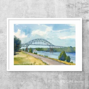 May include: Watercolor painting of a bridge over a river, framed in white. The bridge is a steel arch design, painted in shades of gray. The sky is blue with white clouds, and the water is a deep blue. The Painted Life Studio.