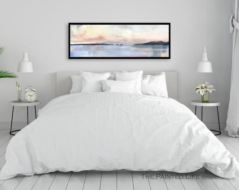May include: A white bed with a white duvet cover and white pillows in a bedroom with a framed abstract watercolor painting of a pink and blue sunset over a body of water. The painting is hanging on a white wall above the bed.