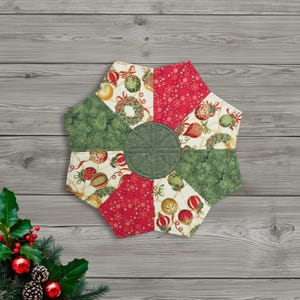 May include: A quilted, hexagonal Christmas-themed trivet. The trivet features a central green circle and is surrounded by segments of red, green, and cream fabric. The segments are decorated with Christmas ornaments, wreaths, and snowflakes. The trivet is approximately 9 inches in diameter.