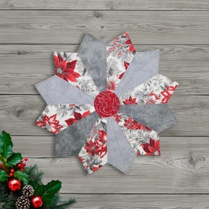 May include: A star-shaped quilted table topper with a festive poinsettia design. The topper features red poinsettias on a white background, gray sections, and a red center. The overall design is approximately 20 inches in diameter.