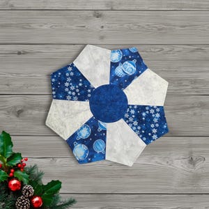 May include: A blue and white quilted Christmas ornament-shaped table topper. The topper features a central blue circle, surrounded by alternating blue patterned sections with snowflakes and ornaments, and white sections with snowflake patterns.