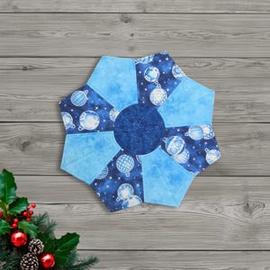 May include: A blue and white quilted Christmas tree skirt with a starburst design. The skirt features a central circle and radiating panels in solid blue and patterned fabric with white ornaments. The overall shape is a six-pointed star.