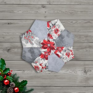 May include: A decorative, hexagonal fabric item featuring a poinsettia design. The item is composed of various fabric pieces in shades of gray, white, and red. The central design showcases large red poinsettia flowers, with gray and white patterned accents.