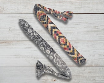 May include: Two fleece dog toys. One is patterned with a geometric design in black, yellow, red, and cream. The other is gray with a white floral pattern. Both have knotted ends.