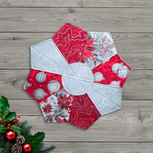 May include: A quilted, hexagonal Christmas-themed table topper. The design features alternating segments of red patterned fabric and light gray fabric. The red segments display patterns of swirls, ornaments, and poinsettias. The center is light gray with stitched lines.