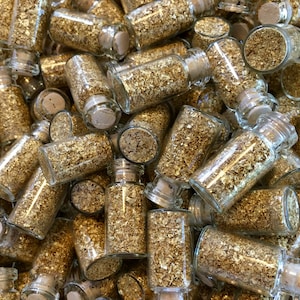 May include: A close-up image of a pile of small glass bottles filled with gold leaf flakes. The bottles have cork stoppers and are arranged in a random pattern.