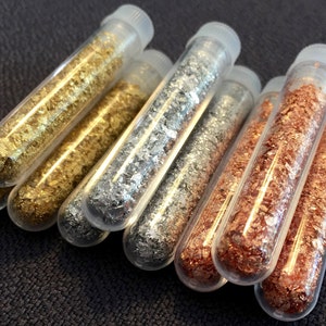 May include: Eight clear glass test tubes filled with metallic flakes. The flakes are gold, silver, and copper.