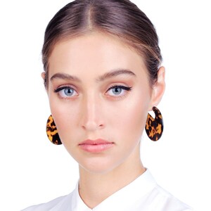 May include: A woman with light brown hair wearing a white collared shirt and large tortoiseshell earrings.