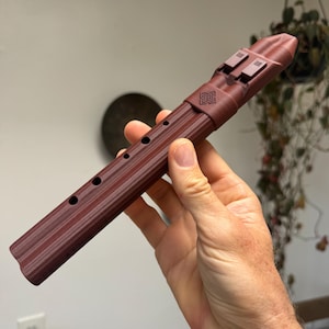 May include: A dark maroon flute with a ridged, cylindrical body featuring five circular finger holes along the side. The instrument has a mouthpiece at the top with two rectangular vents and a small, square-shaped embossed emblem near the head.