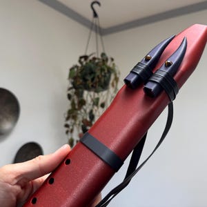 May include: A red ocarina with black accents and a black strap. The ocarina has two dark blue, horn-shaped pieces at the top, secured with black bands. Several holes are visible along the side of the instrument.