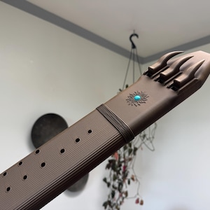 Peut inclure: Une ceinture marron au fini mat avec une série de trous circulaires sur toute la longueur. L'extrémité de la ceinture présente un design à trois branches et est ornée d'un petit emblème solaire circulaire centré avec une pierre turquoise.