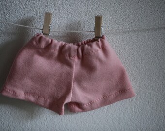 Peut inclure: Un petit short rose pâle suspendu à une corde à linge par deux pinces à linge en bois. Le short est fait d'un tissu doux et possède une taille élastique. L'arrière-plan est un mur uni de couleur claire.
