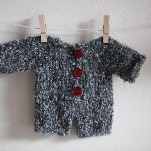 Könnte beinhalten: Ein grau-weiß gestrickter Baby-Overall mit kurzen Ärmeln und Beinen, der mit Holzwäscheklammern an einer Wäscheleine hängt. Der Overall hat drei rote Knöpfe auf der Vorderseite.