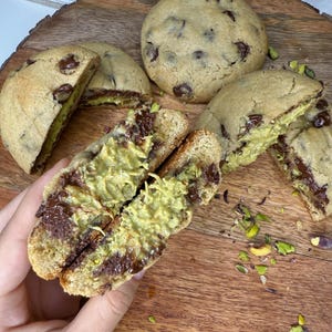 May include: Several chocolate chip cookies, some cut open to reveal a pistachio filling. The cookies are on a wooden board, with scattered pistachio pieces. The cookies have a golden-brown exterior and dark chocolate chips.