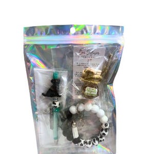 Könnte beinhalten: Eine transparente Plastiktüte mit einem schwarz-weiß gemusterten Armband, einem schwarzen Hundefolie, einem grünen Stift und einer kleinen Glasflasche mit einem grün-weißen Etikett, auf dem "Salted Caramel & Vanilla Bean" steht. Die Tasche enthält auch eine Karte mit dem Text "Anleitung für den Autodiffusor".