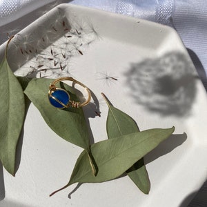 Op de afbeelding: Een goudkleurige, met draad omwikkelde ring met een helderblauwe edelsteen. De ring ligt op een witte zeshoekige schaal, versierd met groene bladeren en delicate zaadhoofdjes. De compositie staat tegen een zachte, witte stoffen achtergrond.