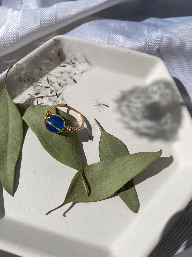 Op de afbeelding: Een goudkleurige, met draad omwikkelde ring met een helderblauwe edelsteen. De ring ligt op een witte zeshoekige schaal, versierd met groene bladeren en delicate zaadhoofdjes. De compositie staat tegen een zachte, witte stoffen achtergrond.