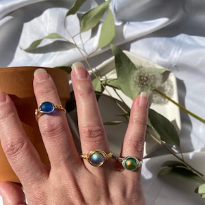 Op de afbeelding: Drie draadgewikkelde ringen met kleurrijke, ronde stenen. De ringen hebben goud- en zilverkleurig draad, met stenen in blauwe, iriserende en groene tinten. De ringen worden op een hand getoond, waarbij het ontwerp en de kleuren van de sieraden worden getoond.