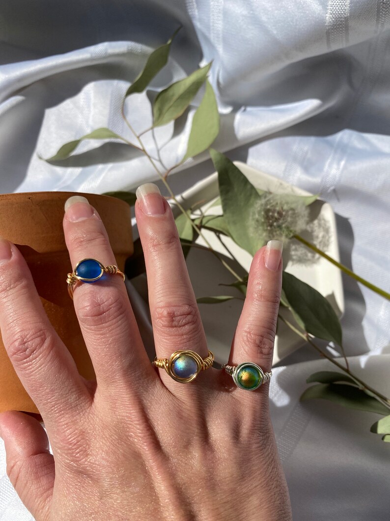 Op de afbeelding: Drie draadgewikkelde ringen met kleurrijke, ronde stenen. De ringen hebben goud- en zilverkleurig draad, met stenen in blauwe, iriserende en groene tinten. De ringen worden op een hand getoond, waarbij het ontwerp en de kleuren van de sieraden worden getoond.