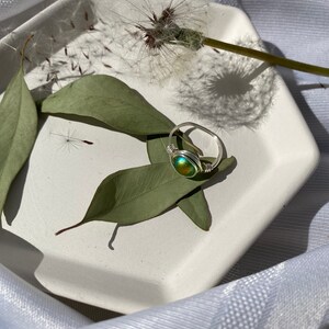 Op de afbeelding: Een handgemaakte ring met draadwikkeling, met een ronde, iriserende steen in groen- en goudtinten. De ring ligt op een witte, geometrische schaal met groene bladeren en een paardenbloem. Een uniek sieraad.