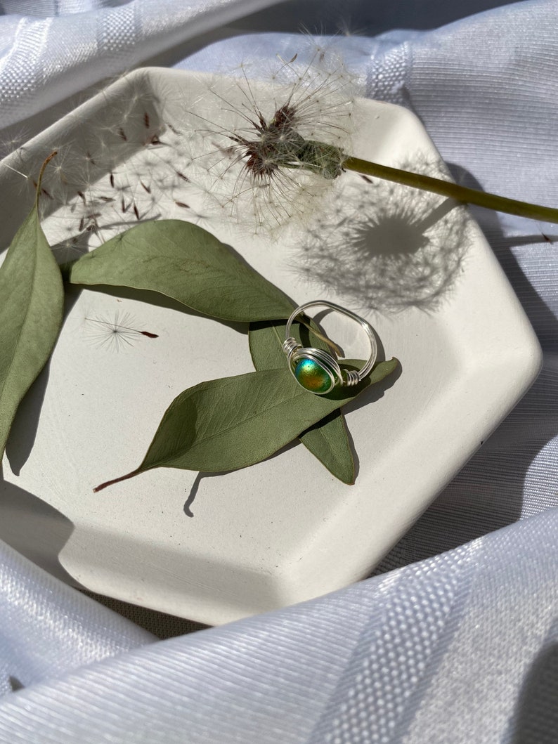 Op de afbeelding: Een handgemaakte ring met draadwikkeling, met een ronde, iriserende steen in groen- en goudtinten. De ring ligt op een witte, geometrische schaal met groene bladeren en een paardenbloem. Een uniek sieraad.