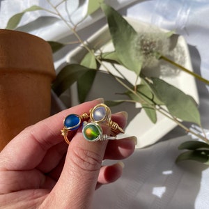 Op de afbeelding: Drie handgemaakte, met draad omwikkelde ringen met kleurrijke glazen kralen. E&eacute;n ring heeft een diepblauwe kraal, een andere een glinsterende iriserende kraal en de derde een lichtpaarse kraal. De ringen worden op een wit oppervlak getoond.