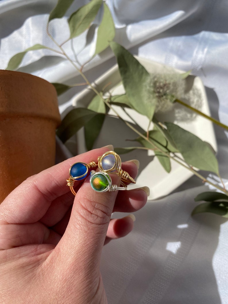Op de afbeelding: Drie handgemaakte, met draad omwikkelde ringen met kleurrijke glazen kralen. E&eacute;n ring heeft een diepblauwe kraal, een andere een glinsterende iriserende kraal en de derde een lichtpaarse kraal. De ringen worden op een wit oppervlak getoond.