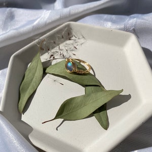 Op de afbeelding: Een goudkleurige ring met een blauwgroene ovale edelsteen, draadomwikkeld, ligt op een witte zeshoekige schaal met groene bladeren. De ring is het middelpunt, tegen een zachte, witte stoffen achtergrond.