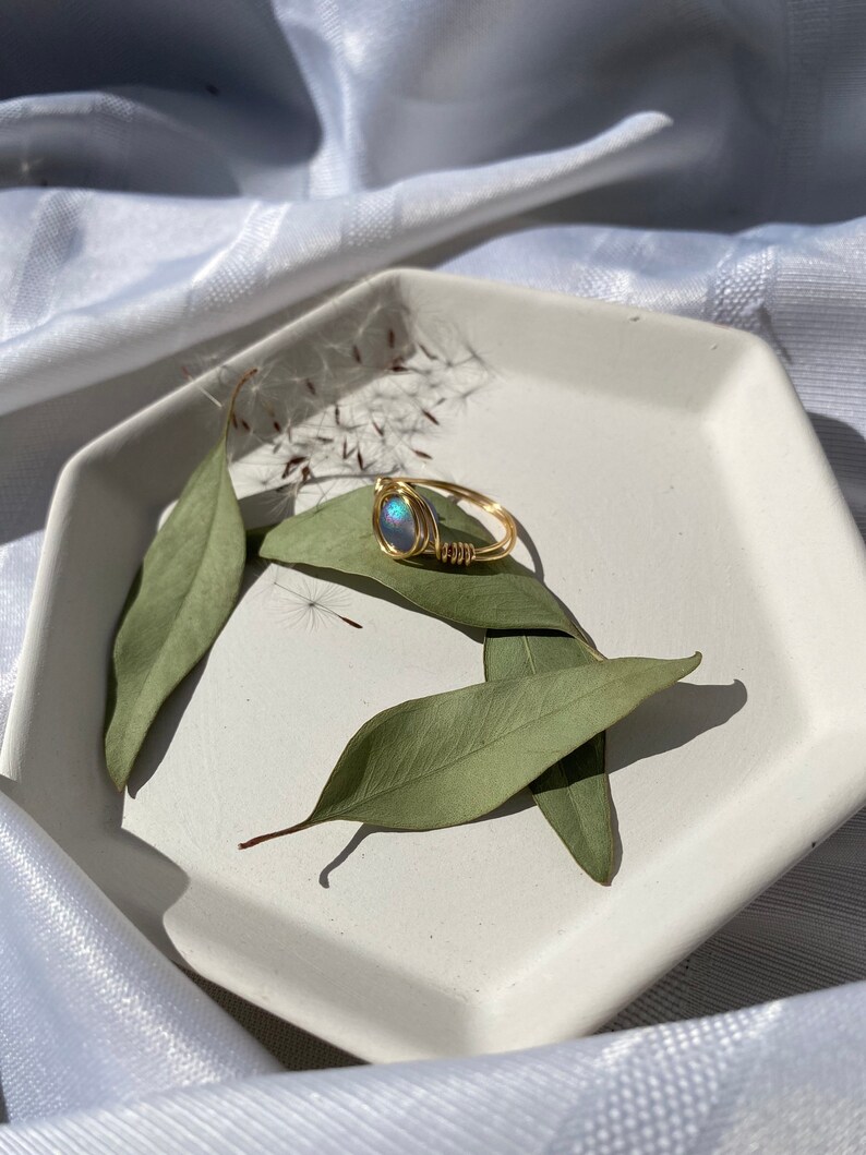 Op de afbeelding: Een goudkleurige ring met een blauwgroene ovale edelsteen, draadomwikkeld, ligt op een witte zeshoekige schaal met groene bladeren. De ring is het middelpunt, tegen een zachte, witte stoffen achtergrond.