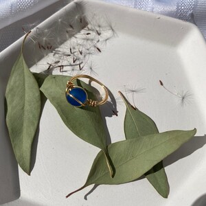 Op de afbeelding: Een goudkleurige, draadgewikkelde ring met een ronde, diepblauwe steen. De ring ligt op een witte vierkante plaat met groene bladeren. Paardenbloemzaden zijn rond de ring verspreid, wat een natuurlijke, aardse uitstraling cre&euml;ert.