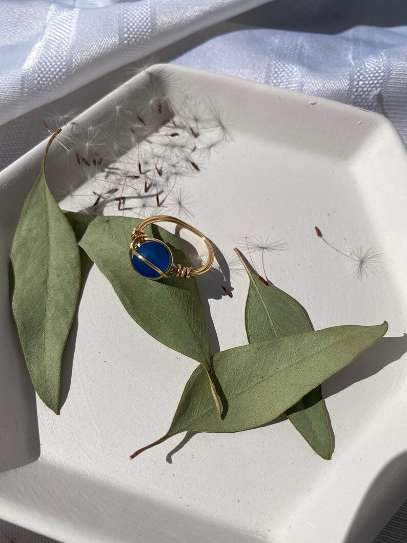 Op de afbeelding: Een goudkleurige, draadgewikkelde ring met een ronde, diepblauwe steen. De ring ligt op een witte vierkante plaat met groene bladeren. Paardenbloemzaden zijn rond de ring verspreid, wat een natuurlijke, aardse uitstraling cre&euml;ert.