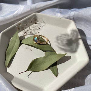Op de afbeelding: Een goudkleurige, draadgewikkelde ring met een blauwgroene ovale steen, rustend op een witte achthoekige schaal met groene bladeren. De ring is het middelpunt en toont het vakmanschap en de iriserende kwaliteit van de steen.