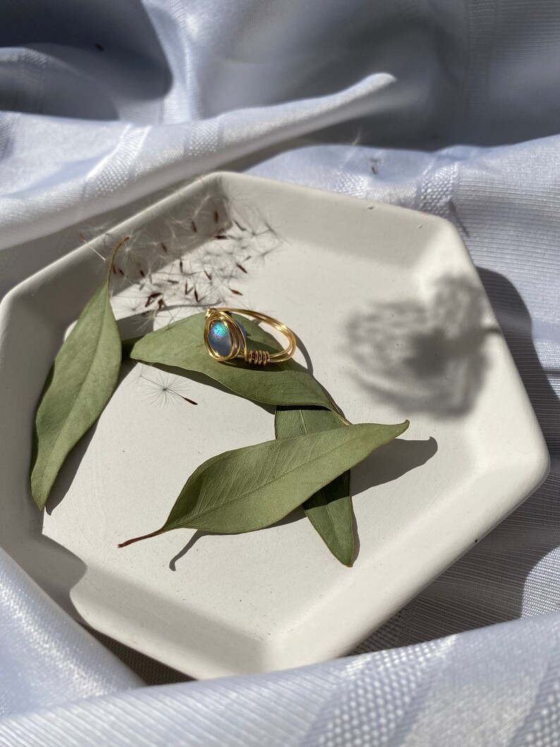 Op de afbeelding: Een goudkleurige, draadgewikkelde ring met een blauwgroene ovale steen, rustend op een witte achthoekige schaal met groene bladeren. De ring is het middelpunt en toont het vakmanschap en de iriserende kwaliteit van de steen.