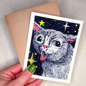 May include: A greeting card featuring a whimsical illustration of a cat with its tongue sticking out, set against a starry night sky. The cat is depicted in shades of gray, with pink accents on its ears and tongue. A brown envelope is included.