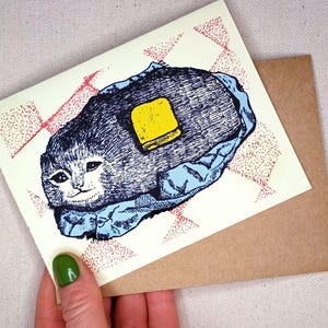 Puede incluir: Una tarjeta de felicitación caprichosa con una ilustración de gato y una nuez de mantequilla. El gato está representado en azul y gris, descansando sobre una manta azul, con un cuadrado de mantequilla amarilla. La tarjeta tiene un patrón de cuadros rojos y blancos y el texto "wyndym baije".