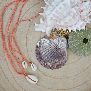 Könnte beinhalten: Eine korallenfarbene Schnurkette mit drei weißen Muschel-Anhängern. Die Kette liegt auf einer Holzoberfläche mit Muscheln und einer kleinen Glasflasche, die mit goldenen Perlen gefüllt ist.
