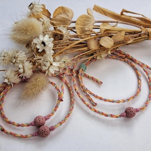 Könnte beinhalten: Geflochtene Armbänder in Pastellfarben mit einer rosa Steinperle. Die Armbänder werden mit einem Trockenblumenstrauß präsentiert. Die Armbänder bestehen aus geflochtenem Kordel in Rosa-, Gelb- und Blautönen.