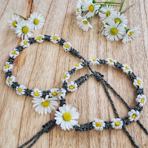 Könnte beinhalten: Zwei graue Freundschaftsarmbänder mit weißen Gänseblümchen, die in die Schnur eingewebt sind. Die Armbänder liegen auf einer Holzoberfläche.