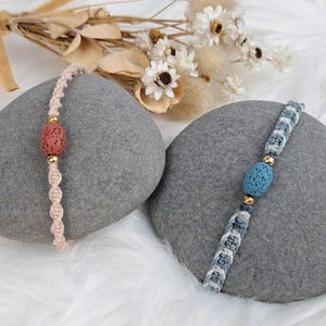 Könnte beinhalten: Zwei Makramee-Armbänder mit Lavasteinperlen. Eines ist hellrosa mit einer korallenfarbenen Perle, das andere blau und weiß mit einer blauen Perle. Beide haben goldfarbene Akzente und werden auf grauen Steinen präsentiert.