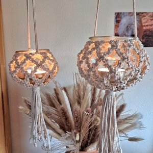 Könnte beinhalten: Zwei Makramee-Kerzenhalter mit Glasbehältern und weißen Quasten, die von der Decke hängen. Die Kerzenhalter sind aus Naturfaserseil gefertigt und haben ein filigranes, kunstvolles Design. Die Behälter sind mit Teelichtern gefüllt, die ein warmes Licht verbreiten.