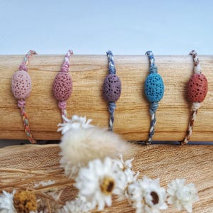 Könnte beinhalten: Fünf handgefertigte Armbänder mit porösen, oval geformten Perlen in Rosa-, Lila- und Blautönen. Die Perlen sind auf geflochtenen Kordeln in komplementären Farben aufgefädelt. Die Armbänder werden auf einem Holzdübel präsentiert.