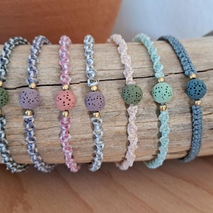 Könnte beinhalten: Sieben handgefertigte Armbänder in verschiedenen Farben. Jedes Armband hat ein geflochtenes Kordeldesign und eine poröse, kugelförmige Perle in Farben wie Grün, Lila und Rosa. Goldfarbene Perlen werden als dekorative Akzente verwendet. Die Armbänder liegen auf einem Stück Treibholz.