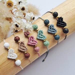 Könnte beinhalten: Eine Sammlung handgefertigter Makramee-Herzarmbänder in verschiedenen Farben, darunter Weiß, Beige, Rosa, Grün, Blau und Schwarz. Jedes Armband hat ein herzförmiges Makramee-Design und ist mit Perlen verziert. Die Armbänder werden auf einem Holzdübel präsentiert.