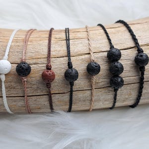 Könnte beinhalten: Eine Sammlung handgefertigter Armbänder mit Lavasteinperlen in verschiedenen Farben. Die Armbänder sind auf unterschiedlich farbigen Kordeln aufgefädelt, darunter Weiß, Schwarz und Braun. Die Lavasteinperlen haben eine poröse Textur.